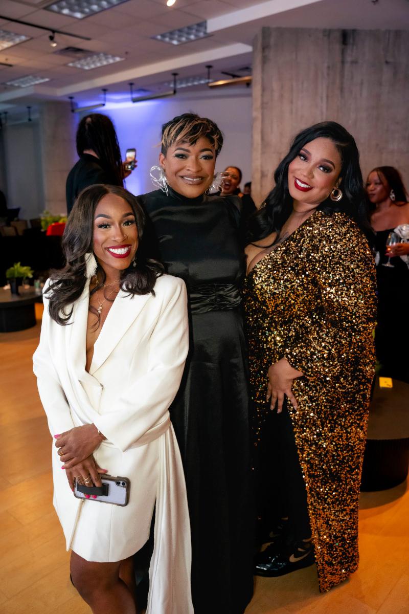 Three women in formal evening attire smile together at an indoor event.