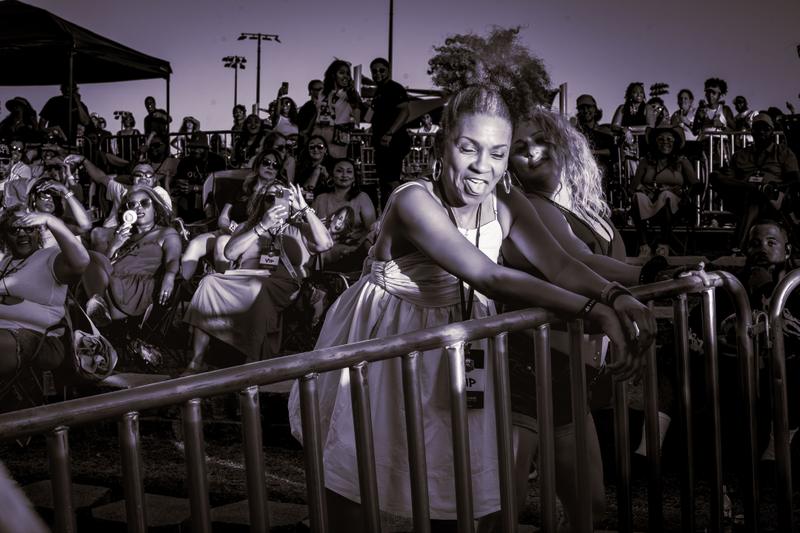 A woman sticking her tongue out while leaning on a metal barricade among a crowd wearing VIP passes at an outdoor event.