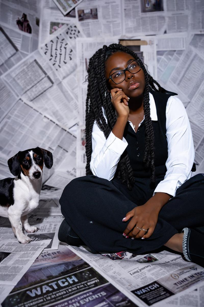 Posed portrait of a young Black woman with glasses and long twists sitting in front of a newspaper backdrop.