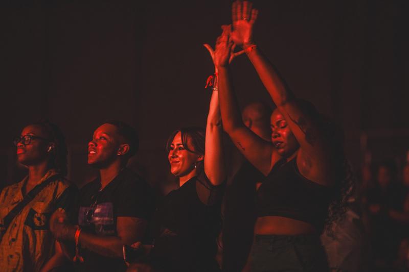 A woman smiling with her arm raised in a crowd under red stage lighting at a concert.