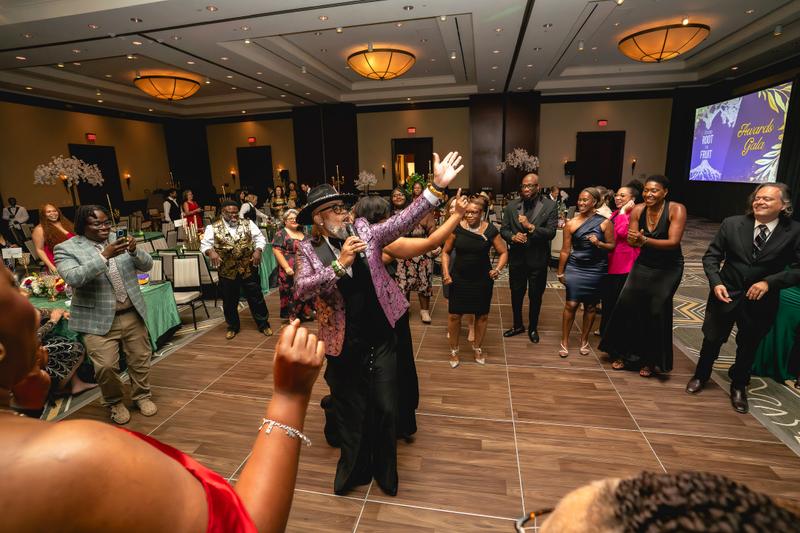 A performer in a pink patterned jacket sings into a microphone surrounded by dancing guests at a formal gala.