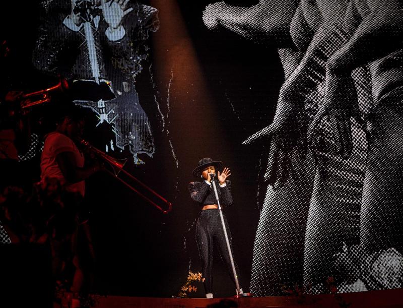 A musician wearing a black sparkling outfit and wide-brimmed hat performs live on stage under a spotlight.