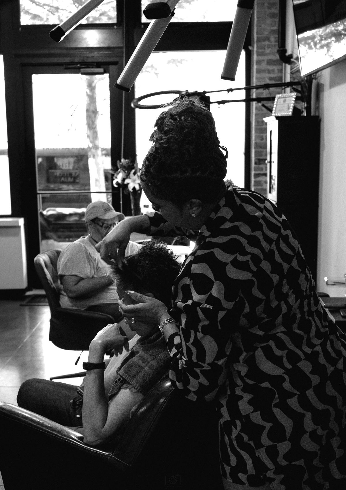 Black and white photo of a hairstylist styling a person's hair under overhead studio lights.