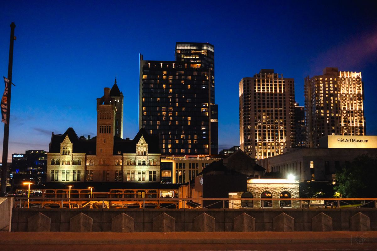Illuminated city buildings and a historic stone structure against a deep blue twilight sky.