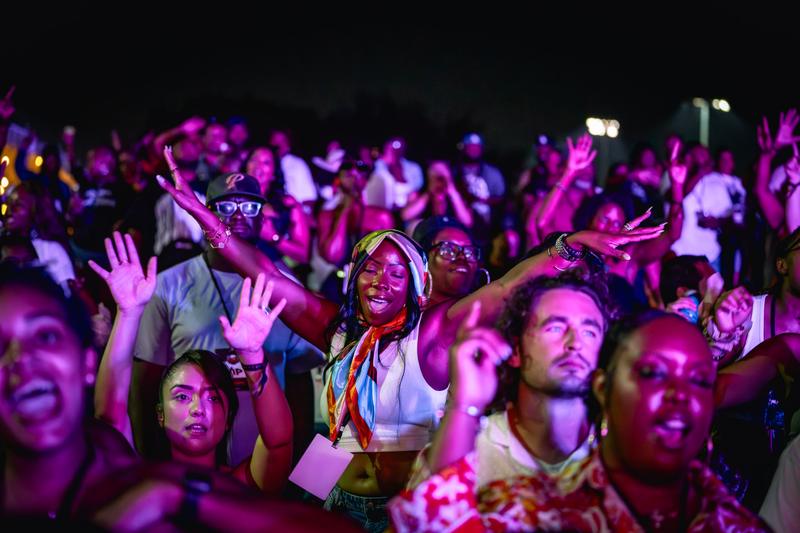 A woman wearing a colorful headscarf dances enthusiastically in a crowd illuminated by vibrant purple stage lights.