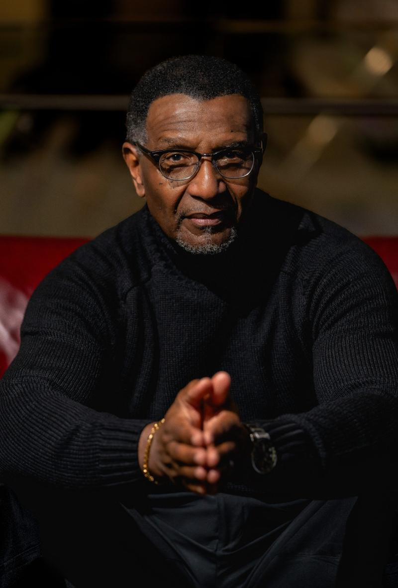 Dramatic portrait of a mature Black man wearing glasses and a black sweater with hands clasped.