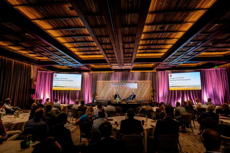 Audience seated at round tables watching two speakers on stage during a Wells Fargo corporate panel discussion.