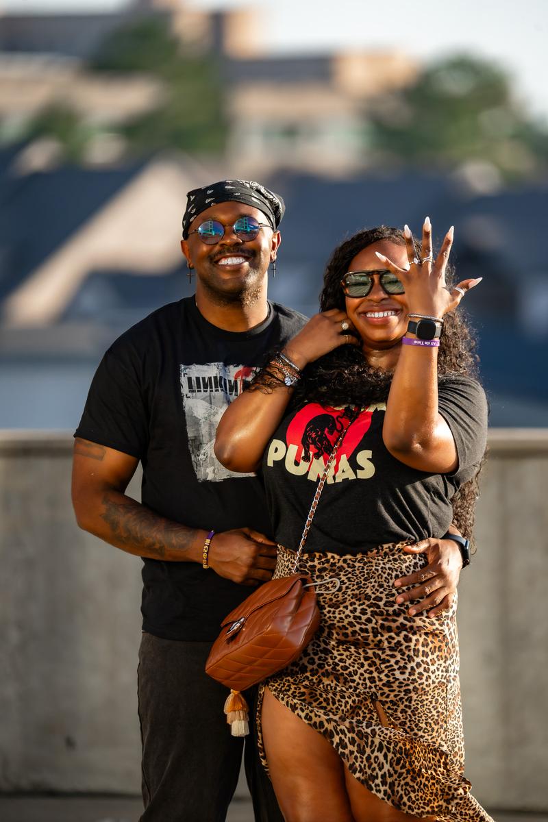 A smiling man wearing a bandana and sunglasses poses outdoors with a woman wearing a graphic t-shirt and leopard print skirt.