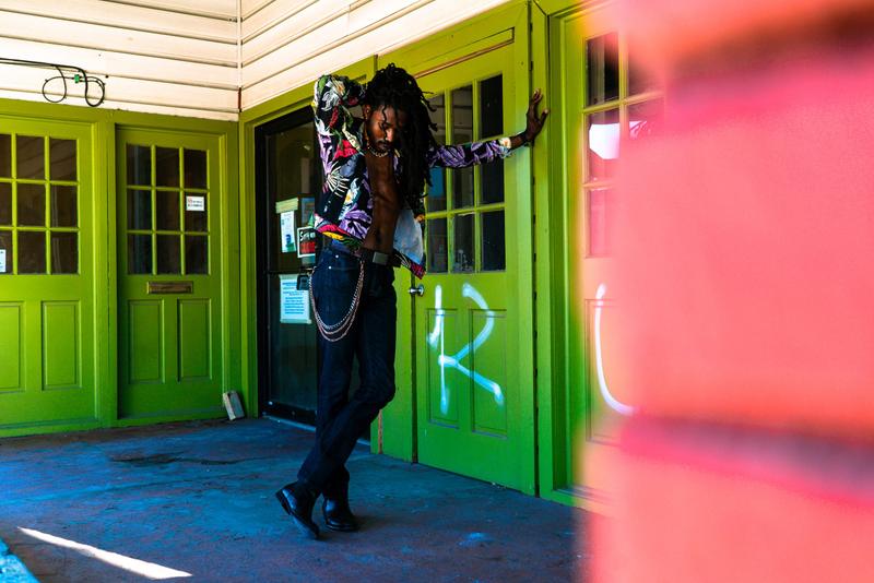 A man with long dreadlocks and an unbuttoned floral jacket leaning against a bright green door.