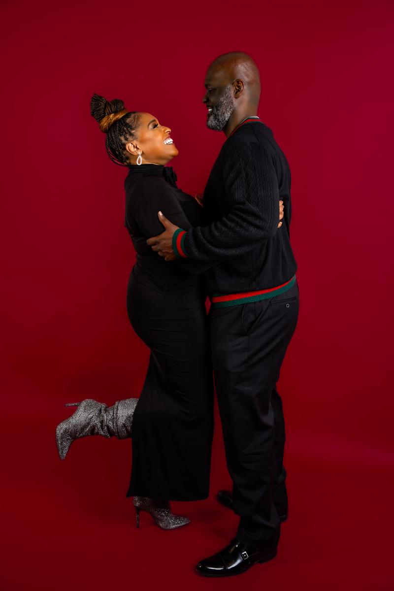A smiling couple dressed in black posing together against a solid dark red studio background.