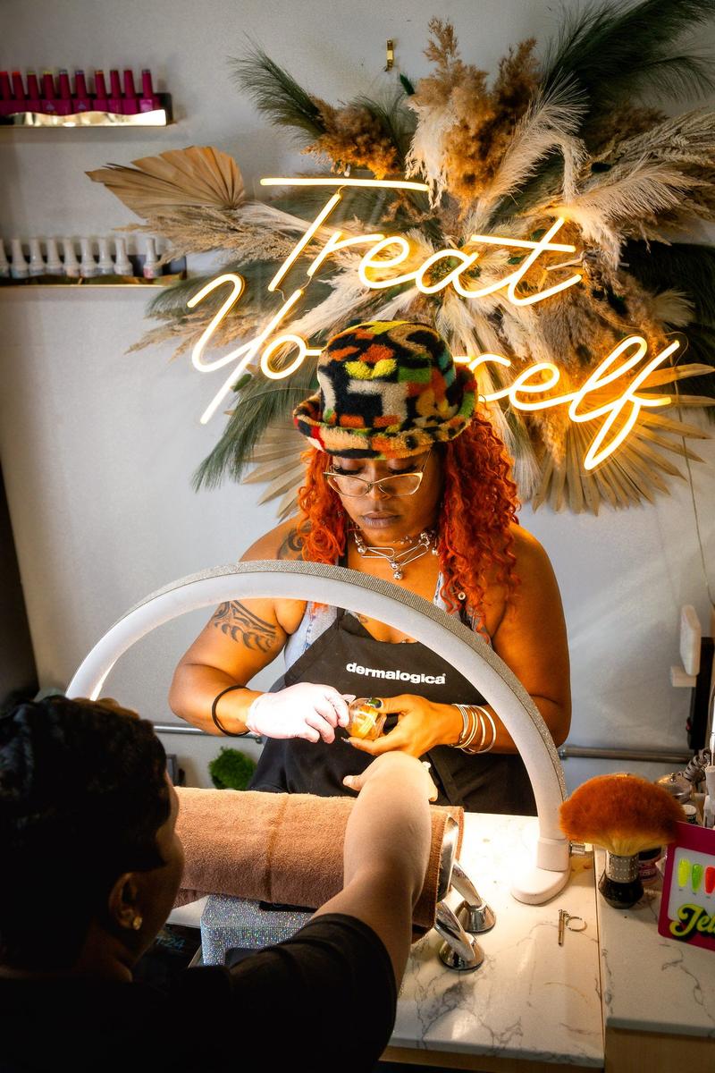 A beauty professional wearing a colorful hat and Dermalogica apron works on a client under a ring light.