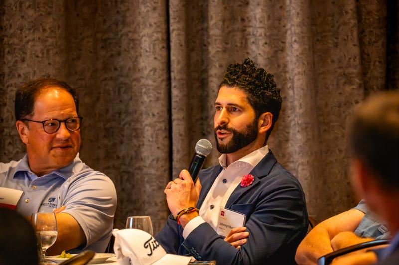 A bearded man wearing a suit jacket and name badge speaks into a microphone at a corporate event.