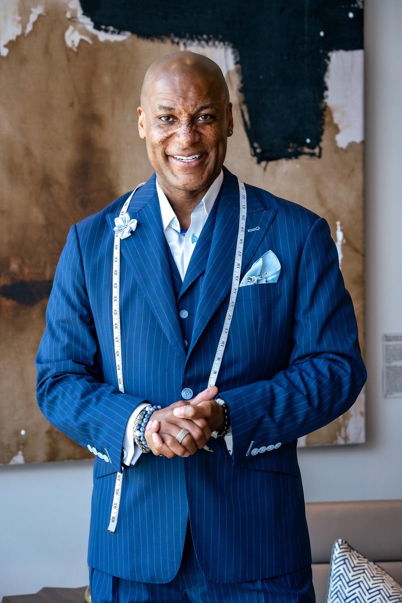 Portrait of a smiling man wearing a blue pinstripe suit with a tailor's measuring tape draped around his neck.