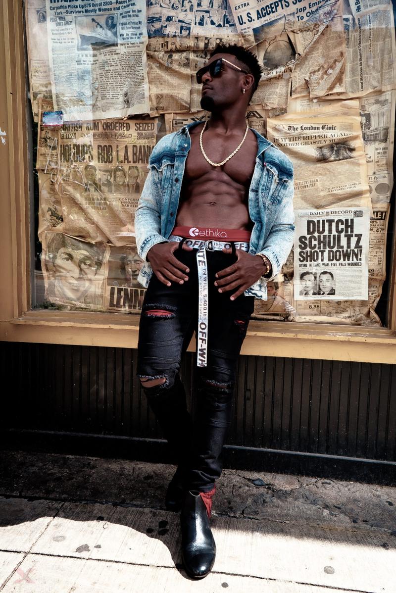 A man in an open denim jacket, sunglasses, and black distressed jeans poses in front of a newspaper display window.