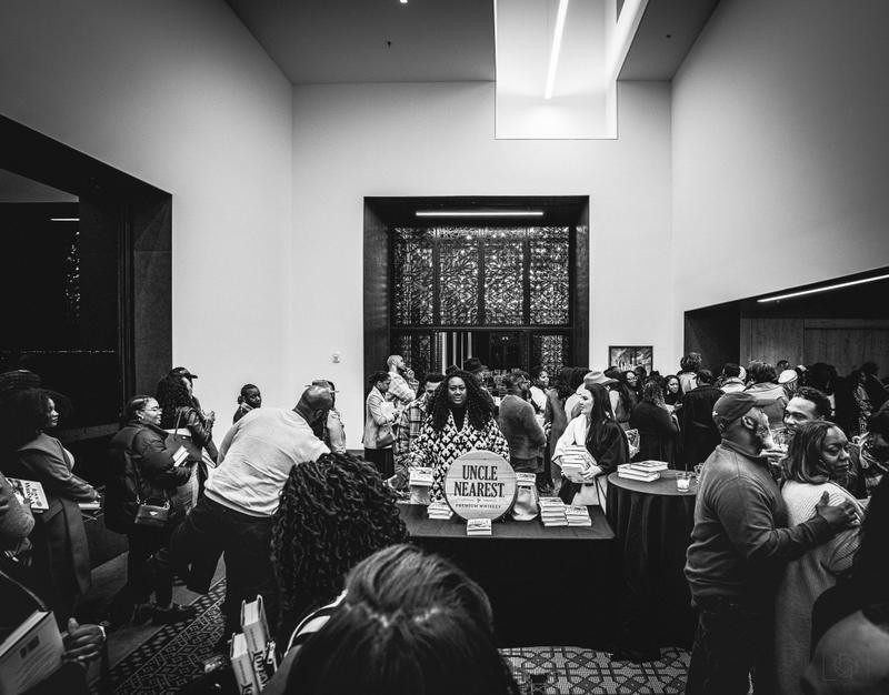 Black and white photo of a woman standing behind an Uncle Nearest Whiskey table at a crowded indoor event.