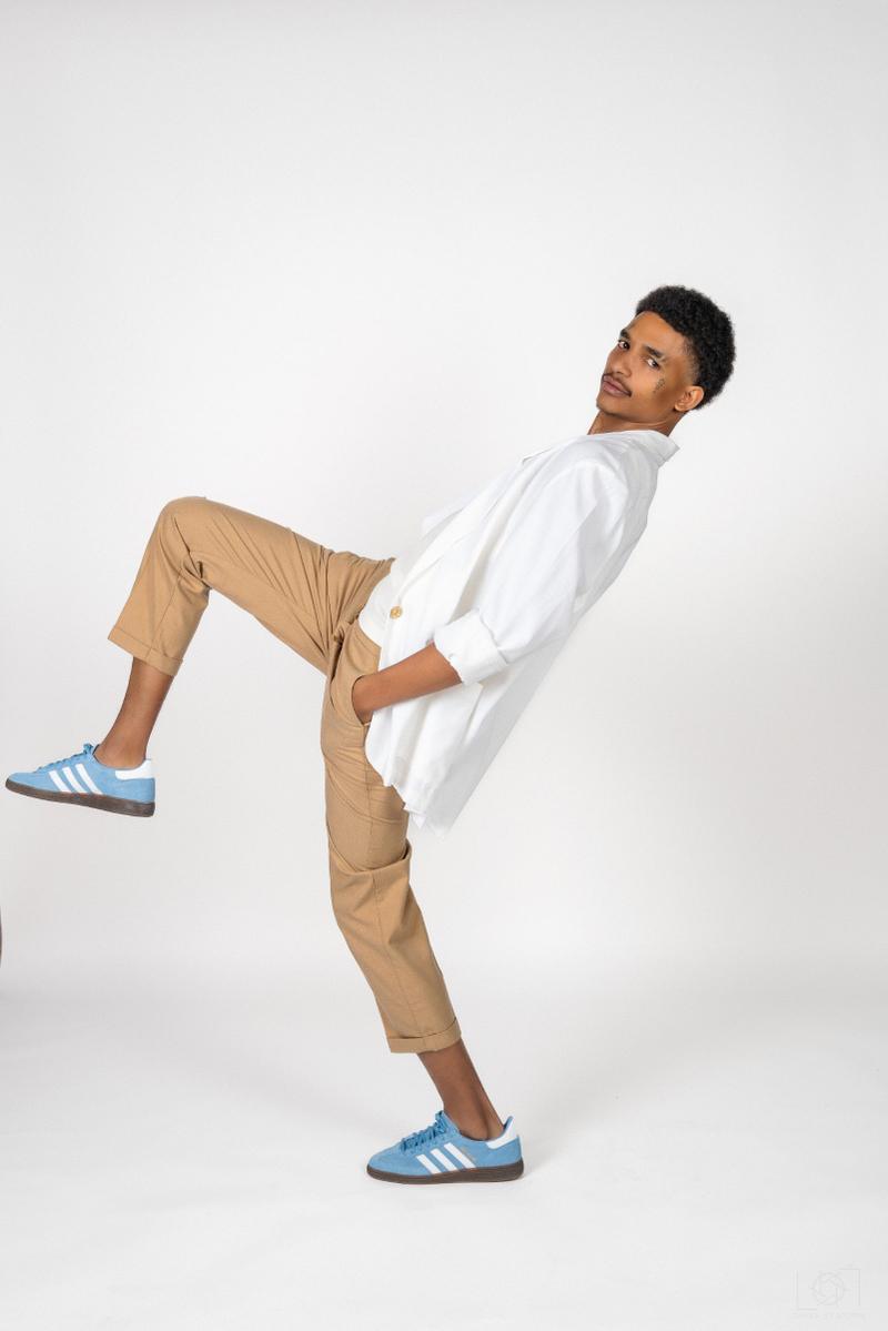 A man in a white jacket, tan pants, and blue sneakers poses dynamically in a studio shoot against a white backdrop.