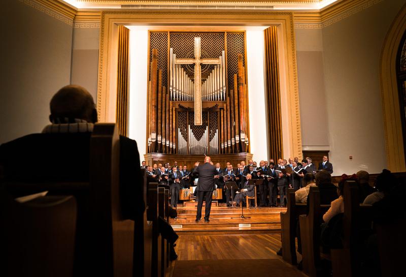 A conductor directs a choir performing on stage in front of a large pipe organ and cross inside a church sanctuary.