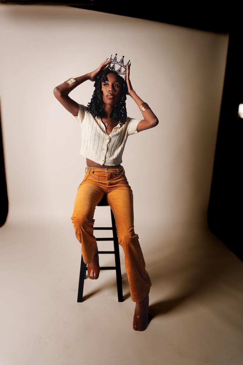 A woman wearing a crown, cream top, and mustard pants poses on a stool in a studio shoot.