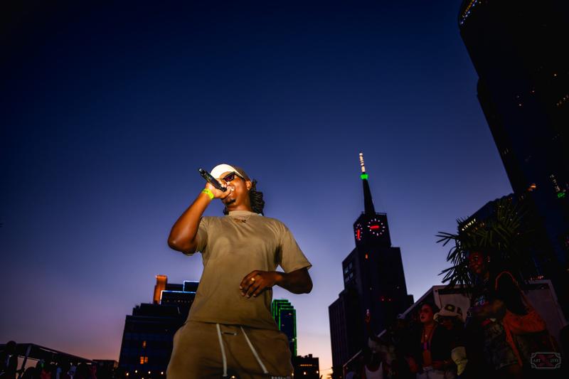 A musician performs with a microphone outdoors at dusk with the Dallas skyline in the background.