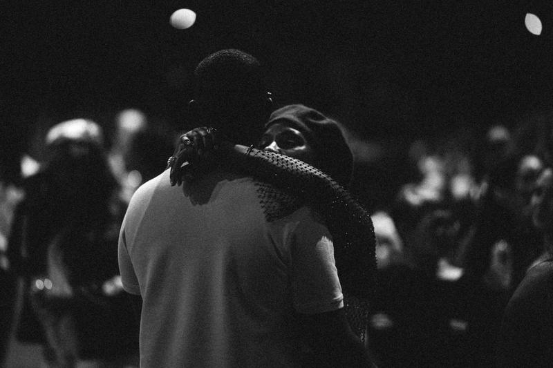 Black and white shot of a musical artist wearing a beret hugging someone under dark stage lighting.