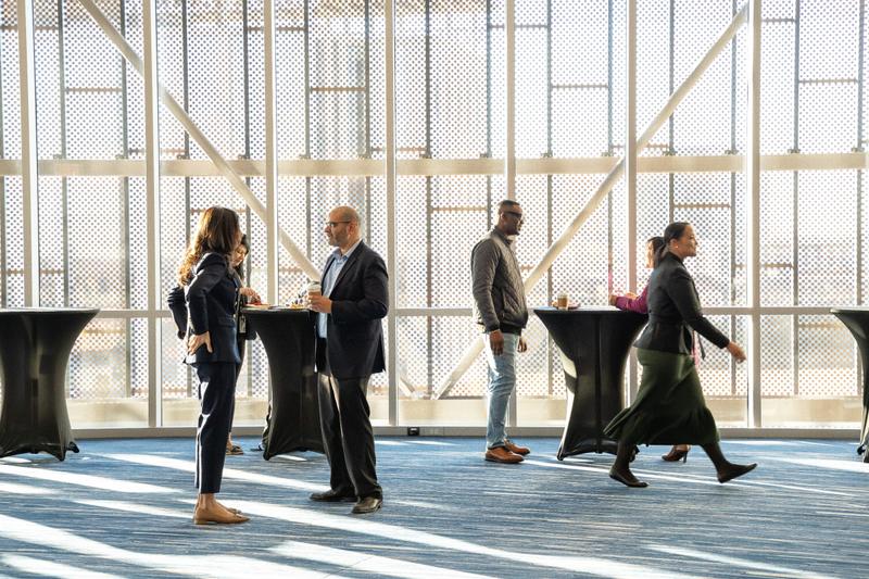 Two men stand near large patterned windows holding coffee cups during a corporate event break.