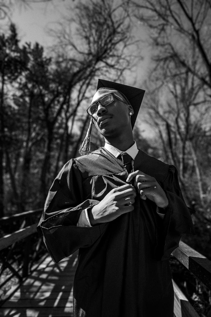 Black and white low-angle portrait of a young man wearing a graduation cap and gown outdoors.