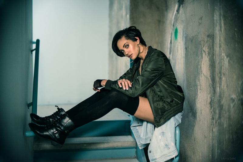 A woman with short dark hair, hoop earrings, and an olive bomber jacket poses while sitting on concrete stairs.