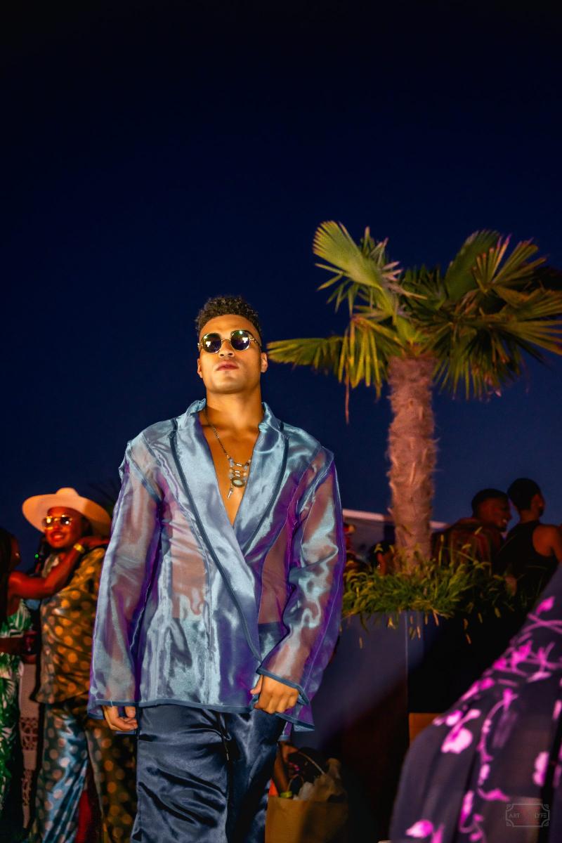 A male model wearing a sheer blue jacket and round sunglasses walks down an outdoor runway at night.