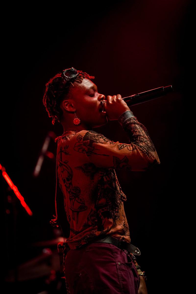 A musical performer singing into a microphone under red stage lighting.