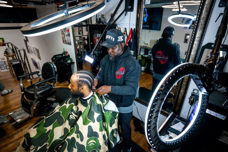 A barber wearing a black hoodie adjusts a haircut cape for a client inside a barbershop with ring lights.