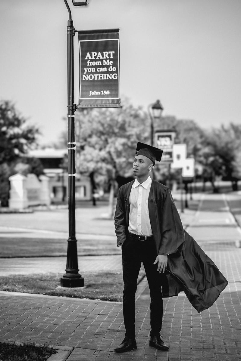 Black and white portrait of a graduate in a cap and gown posing next to a lamp post outdoors.