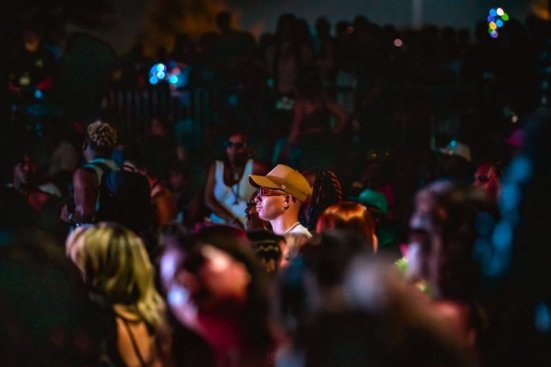 Side profile of a person wearing a cap and sunglasses in a dimly lit crowd at a concert event.