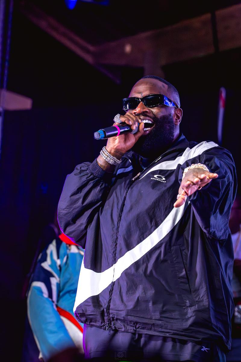 Rapper Rick Ross performing live on stage into a microphone while wearing sunglasses.