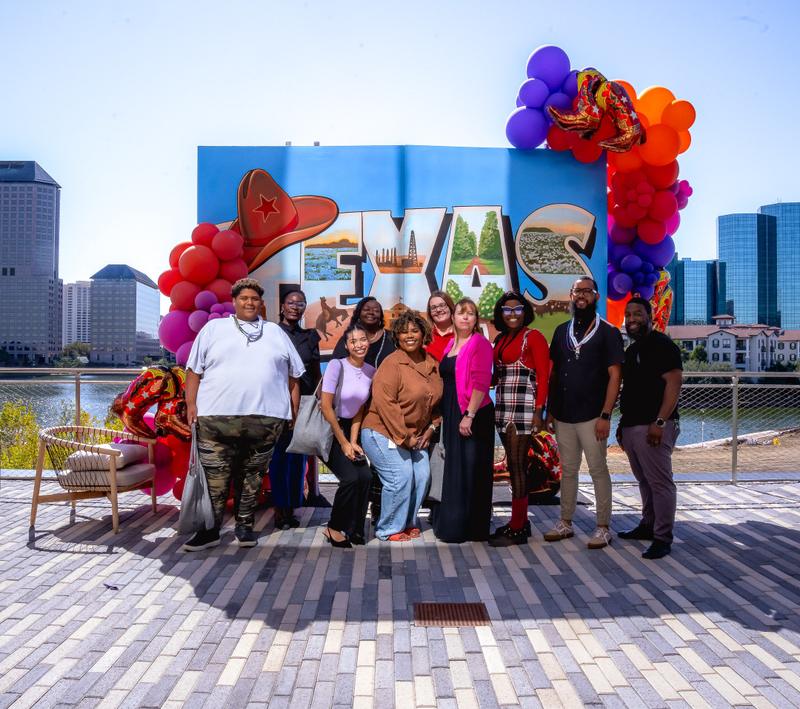 A group of women pose together smiling in front of a large Texas mural at an outdoor event.