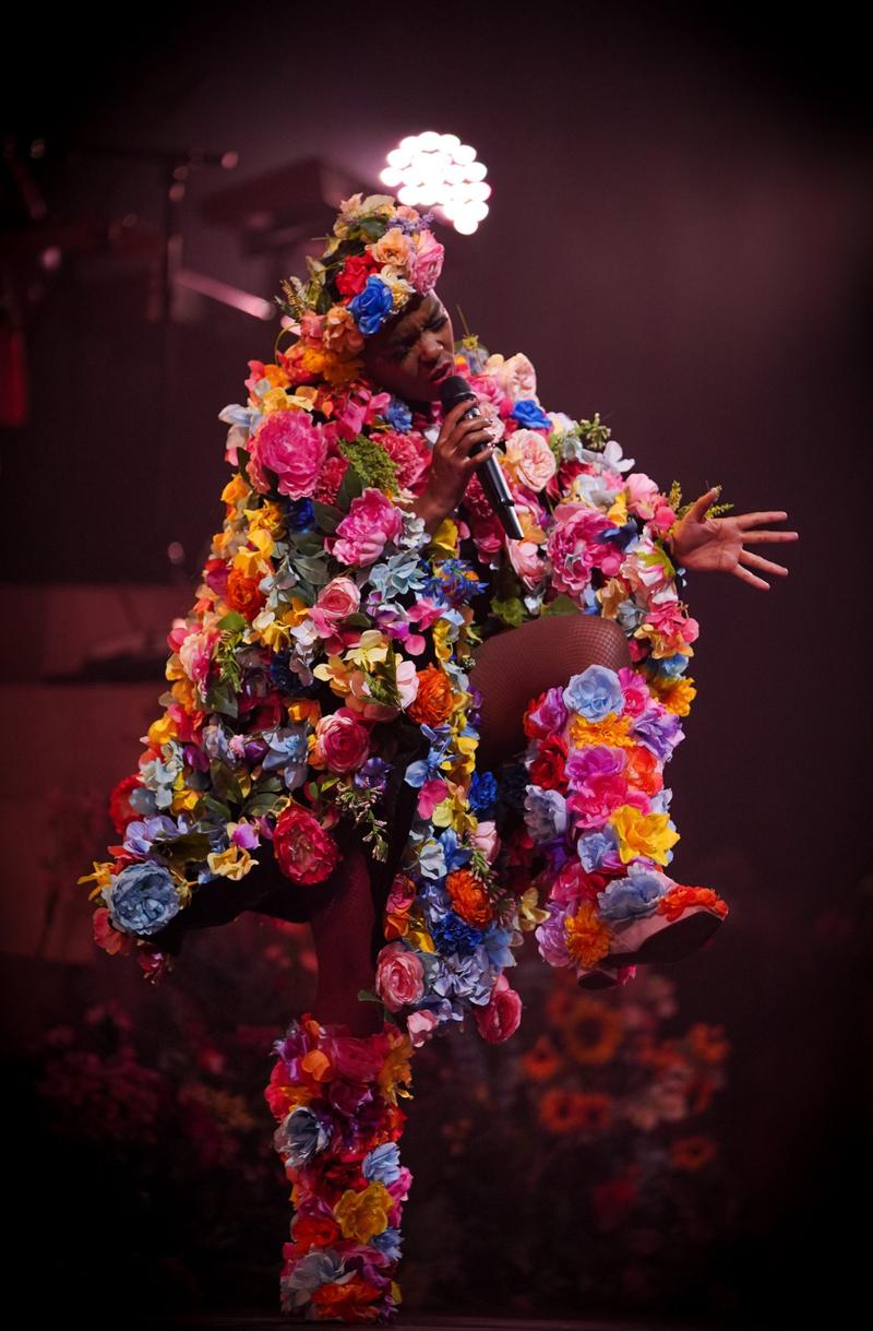 A singer wearing an elaborate suit covered in colorful flowers performs on stage into a microphone.