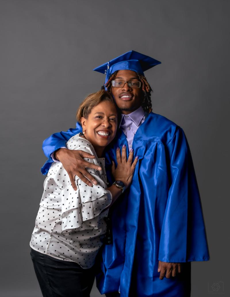 A smiling young male graduate in a blue cap and gown hugging a woman wearing a polka dot top.