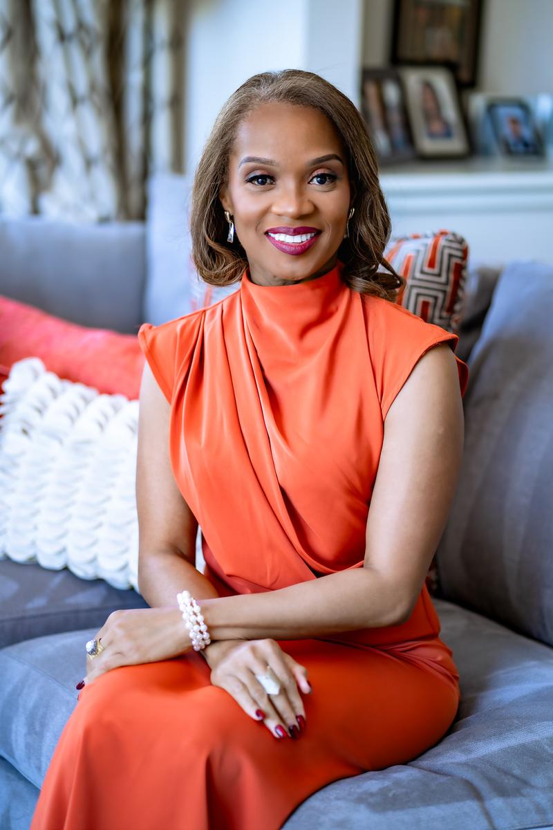 Portrait of a smiling woman in an orange high-neck top sitting indoors.