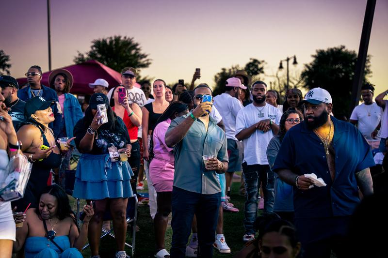 A man in sunglasses holds up his smartphone to record while standing in an outdoor crowd at sunset.