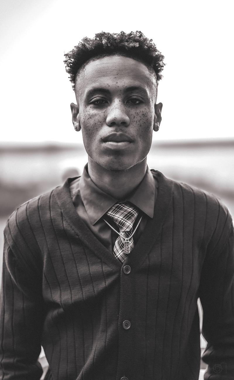 Black and white portrait of a young man with freckles wearing a collared shirt, plaid tie, and cardigan sweater.