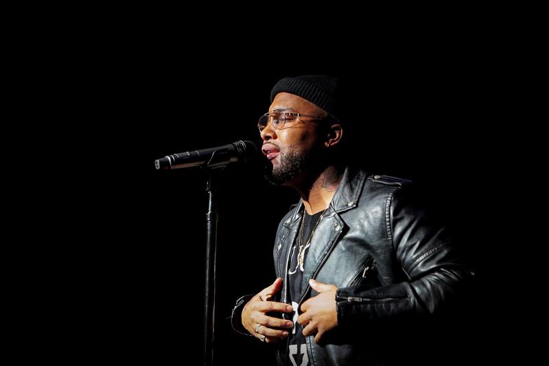 Musician wearing a black beanie and leather jacket singing into a microphone on stage.