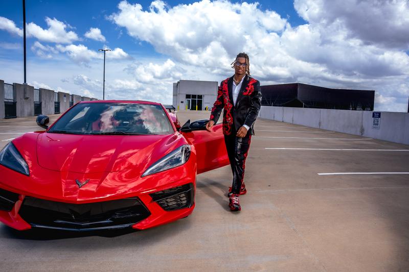 A young man in a black and red embellished suit poses next to a red sports car on a parking garage roof under a cloudy sky.