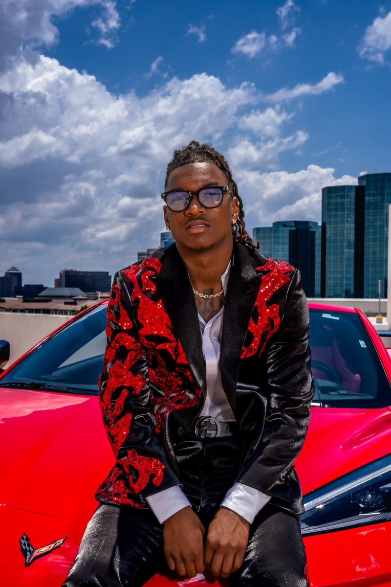 Posed outdoor portrait of a man wearing a black suit jacket with red sequined embroidery and glasses against a bright blue sky.