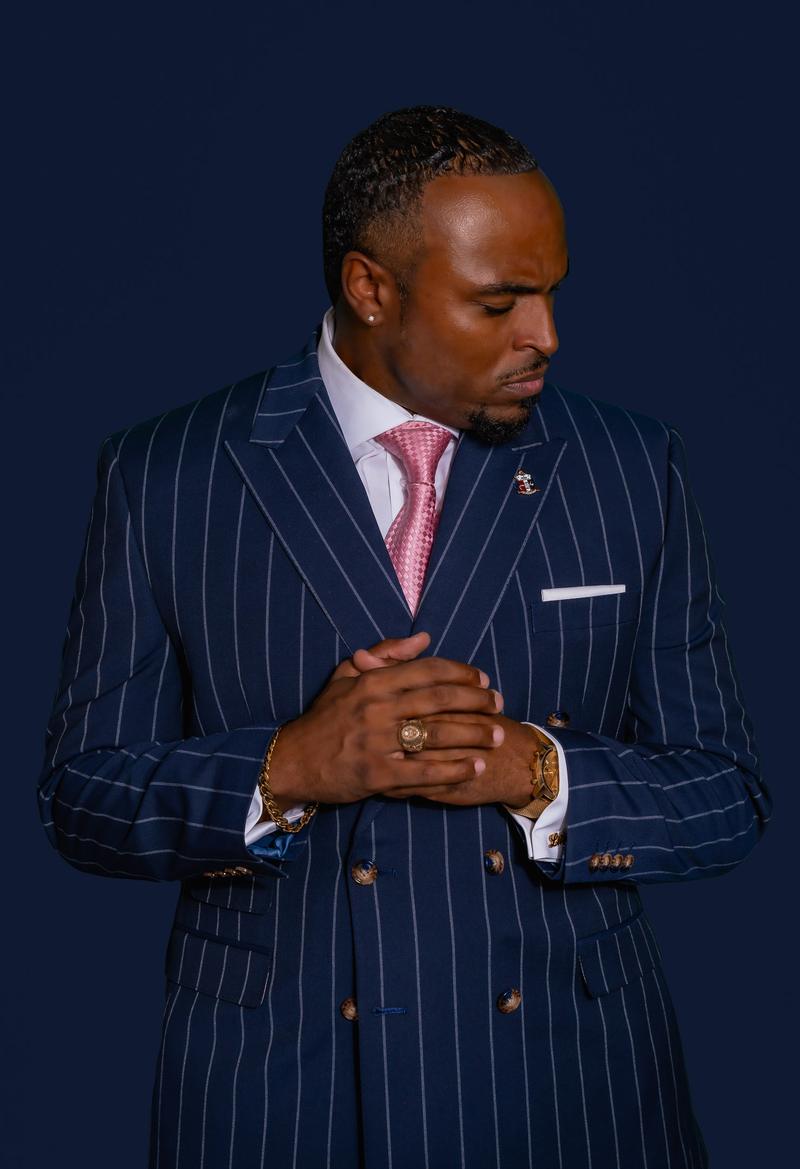 Man in a blue pinstripe suit and pink tie posing with clasped hands against a dark background.