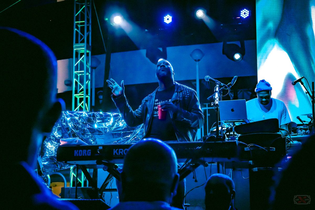 A musician on stage under blue spotlights gesturing with one hand raised while standing behind a keyboard synthesizer.