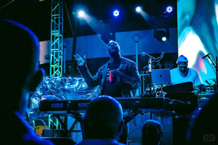 A musician on stage under blue spotlights gesturing with one hand raised while standing behind a keyboard synthesizer.