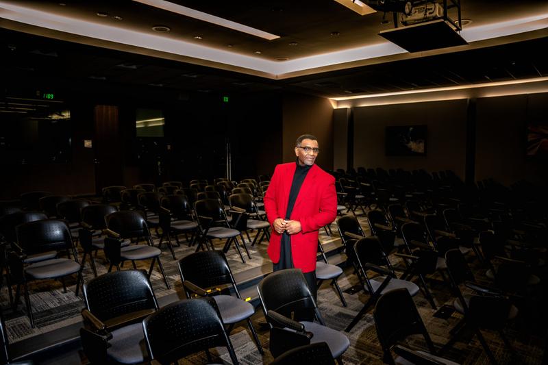 A man in a red blazer and glasses poses for a portrait in an empty conference auditorium with rows of chairs.