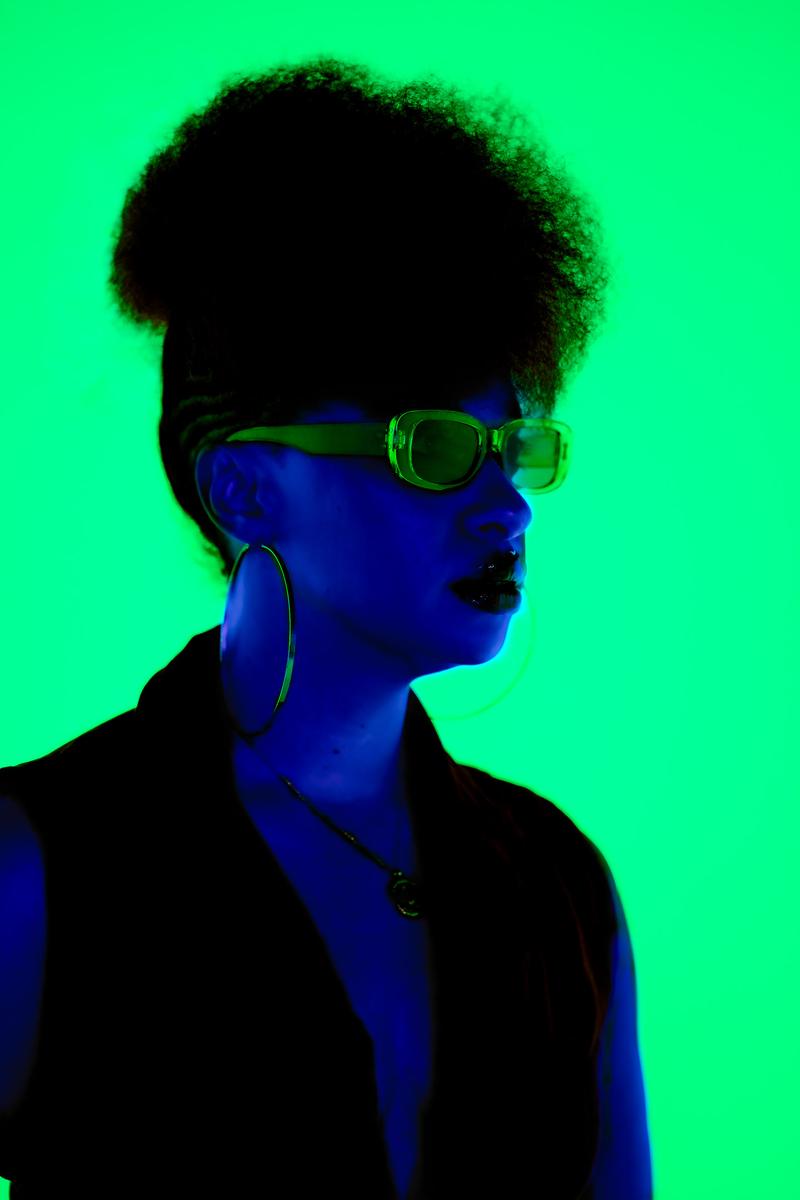 Styled portrait of a person with an afro and sunglasses lit in vivid blue and green neon tones.