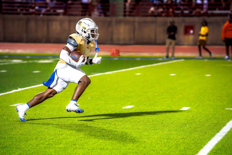 American football player in a helmet and gold jersey running with the ball on a turf field.