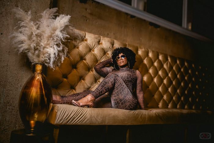 A woman in sunglasses and a patterned jumpsuit reclines on a tufted gold velvet bench beside a tall vase of pampas grass.