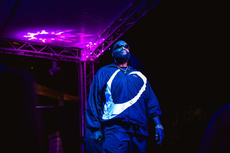 A male musical artist wearing sunglasses and a track jacket stands under blue and purple concert stage lighting.